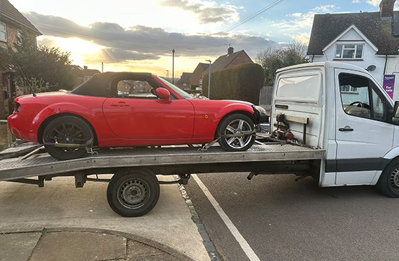 red sports car being towed on flatbed truck with sunset backdrop and residential area nearby