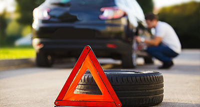 a person changing a tire on a car with a red warning triangle in the foreground showing car maintenance safety tips and assistance in case of emergencies