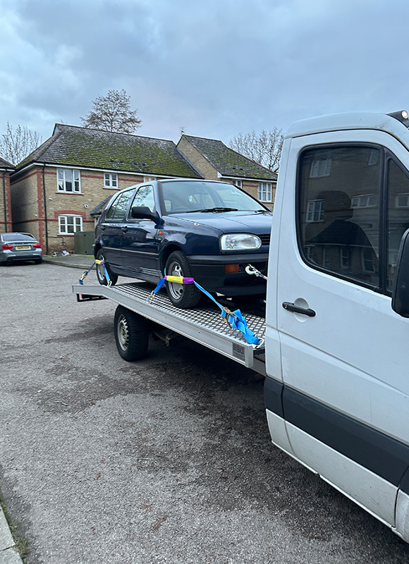 tow truck transporting a blue car on a flatbed with secure straps in a residential area featuring eight trees in the background